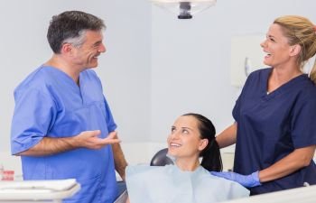 A dentist and his assistant talking to a female patient sitting in a dental chair.