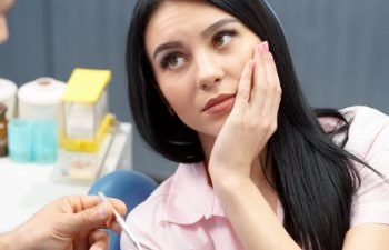 A black-haired woman with dental pain sitting in a dental chair and holding her cheek when discussing the issue with the dentist.