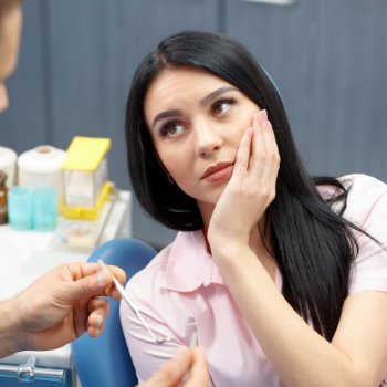 A black-haired woman with dental pain sitting in a dental chair and holding her cheek when discussing the issue with the dentist.