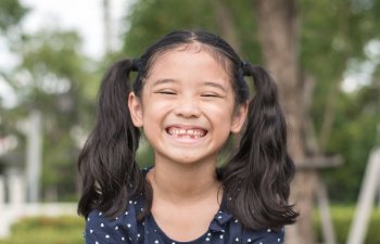 A young girl with black pigtails and a wide smile shows missing front teeth. She is wearing a navy blue polka dot shirt and is outdoors with green trees in the blurred background.